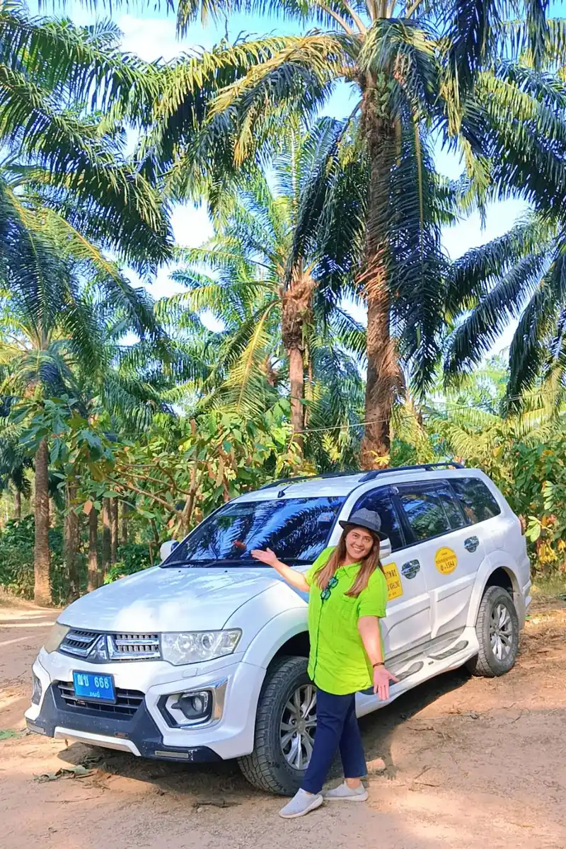 Smiling driver next to a white SUV taxi under oil palm trees near Krabi